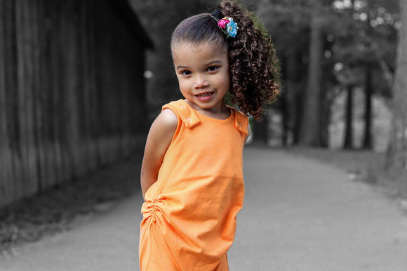 Afro Colombian child outside standing with hand behind their back, smiling. Tight Curly hair in high side pony tail, blue and pink flowers at base of pony tail.  Child is wearing an orange tank style romper With hidden torso adjusters and shoulder snaps.