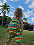 Blonde Caucasian child, hair covering face, standing outside in grass with palm trees and clouds behind them wearing a knee length bright rainbow striped tank romper with torso adjusters, shoulder snaps, Kangaroo pocket, medipocket with a snap and inseam snaps