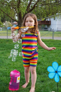Brown haired Caucasian girl blowing bubbles outside in the grass in front of a tree wearing a rainbow stripe short length tank romper with hidden torso adjusters and shoulder snaps.